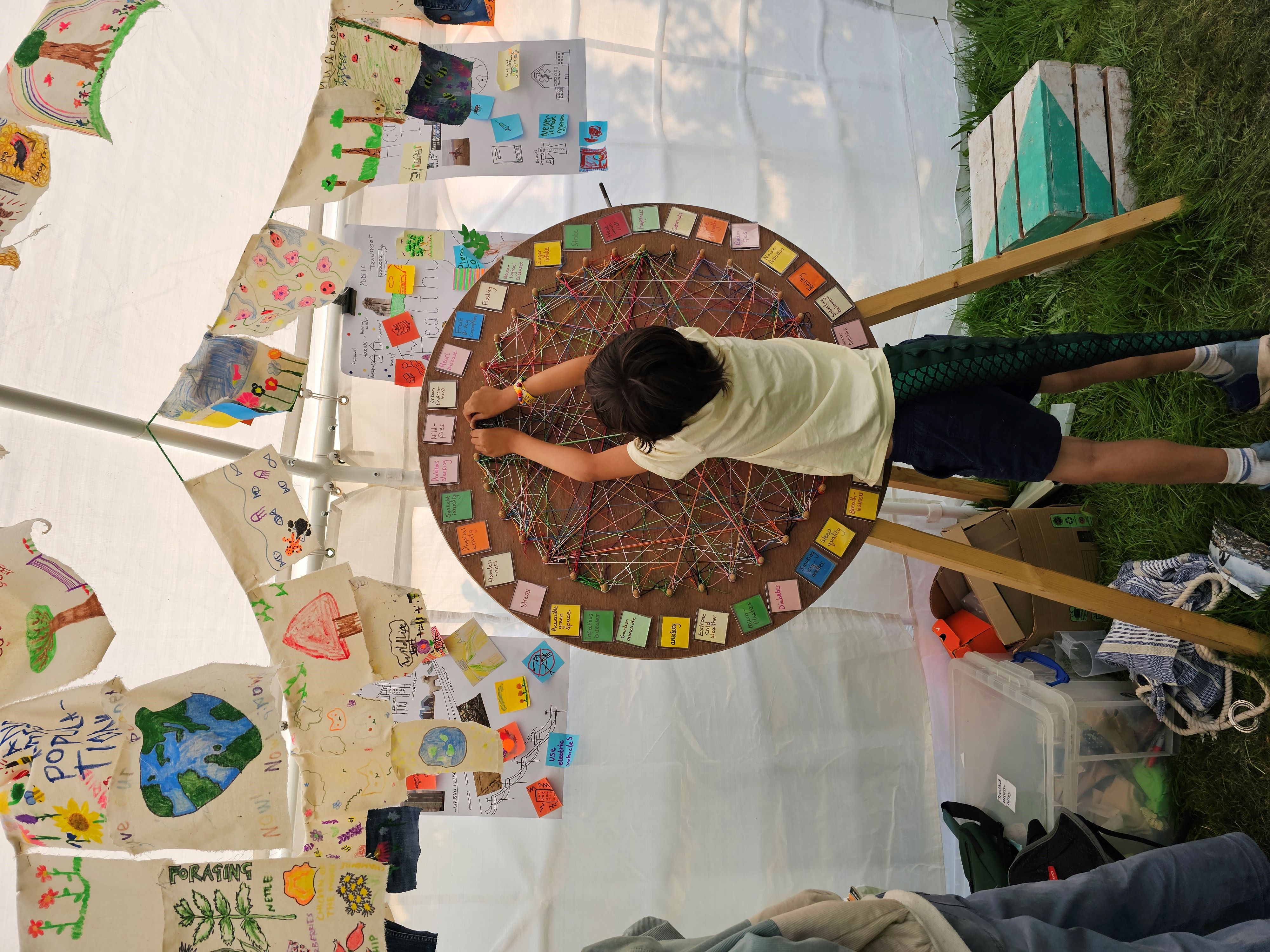 A young boy stretching up in front of a large wooden wheel covered in colourful strings. Overhead is hand-decorated bunting.